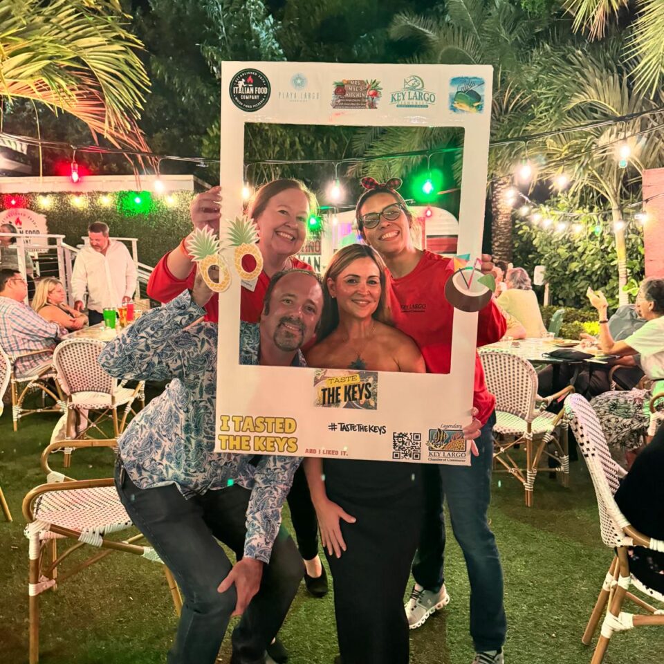 Group posing with a Taste the Keys photo frame on a lawn under string lights