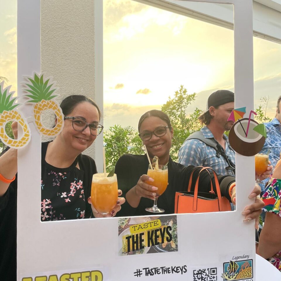Guests with drinks posing in an I Tasted the Keys photo frame at sunset